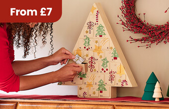 A person putting a small gift into a personalised wooden tree advent calendar. From &pound;7 badge. Clicking leads to advent calendar products.